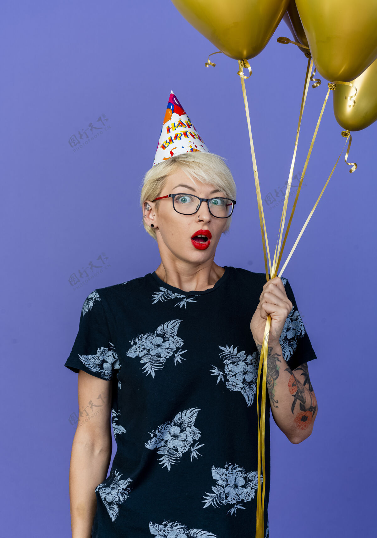 印象令人印象深刻的年轻金发党妇女戴着眼镜和生日帽手持气球看着前面孤立的紫色墙壁年轻气球衣服