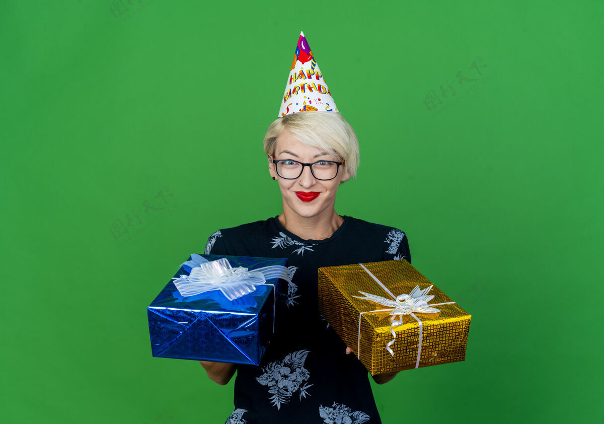 眼镜年轻的金发派对女郎戴着眼镜 戴着生日帽 手里拿着礼盒 看着前面隔离的绿色墙壁上的复制空间礼物脸人