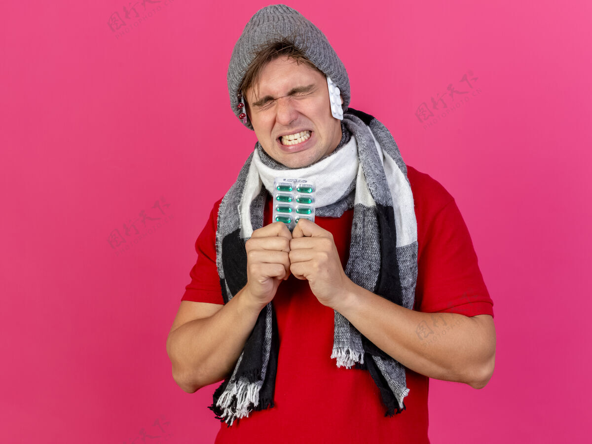 围巾年轻英俊的金发病患 戴着冬天的帽子 戴着围巾 抱着一包药 闭着眼睛 帽子下有一包药 隔离在粉红色的墙上 留着复印空间疾病表情人