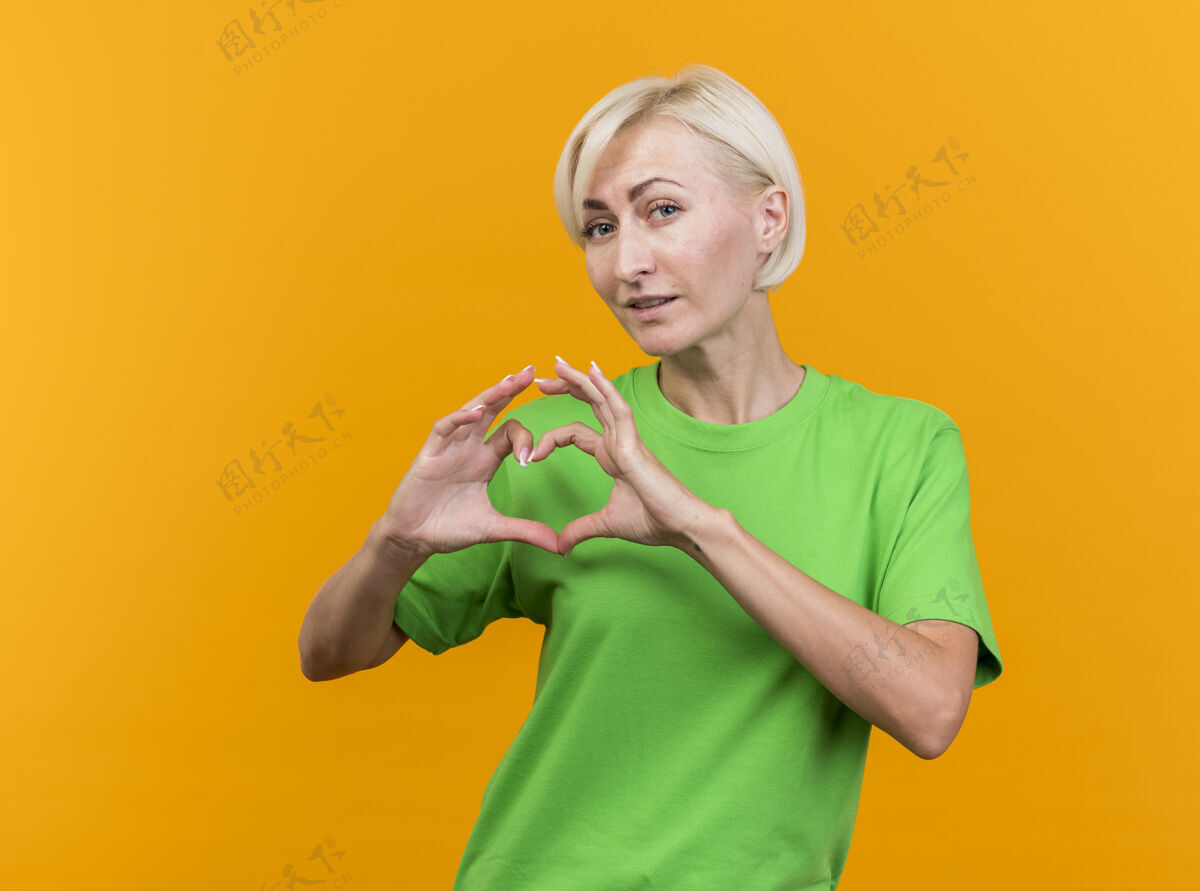 人自信的中年金发斯拉夫女人看着前面做心脏标志隔离在黄色的墙壁与复制空间自信立场女人