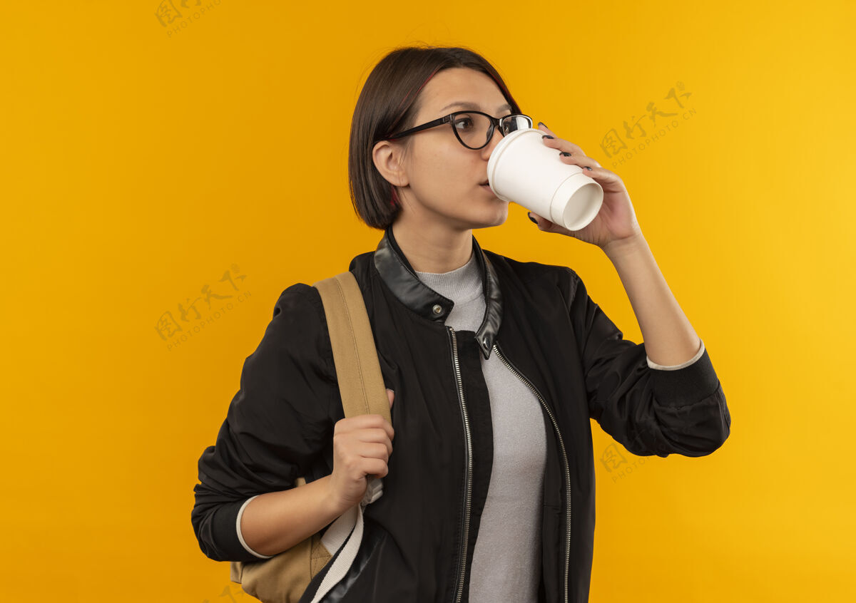 年轻年轻的女学生戴着眼镜 背着咖啡袋从塑料咖啡杯里喝咖啡 一边看着孤立的橙色背景杯子女孩穿