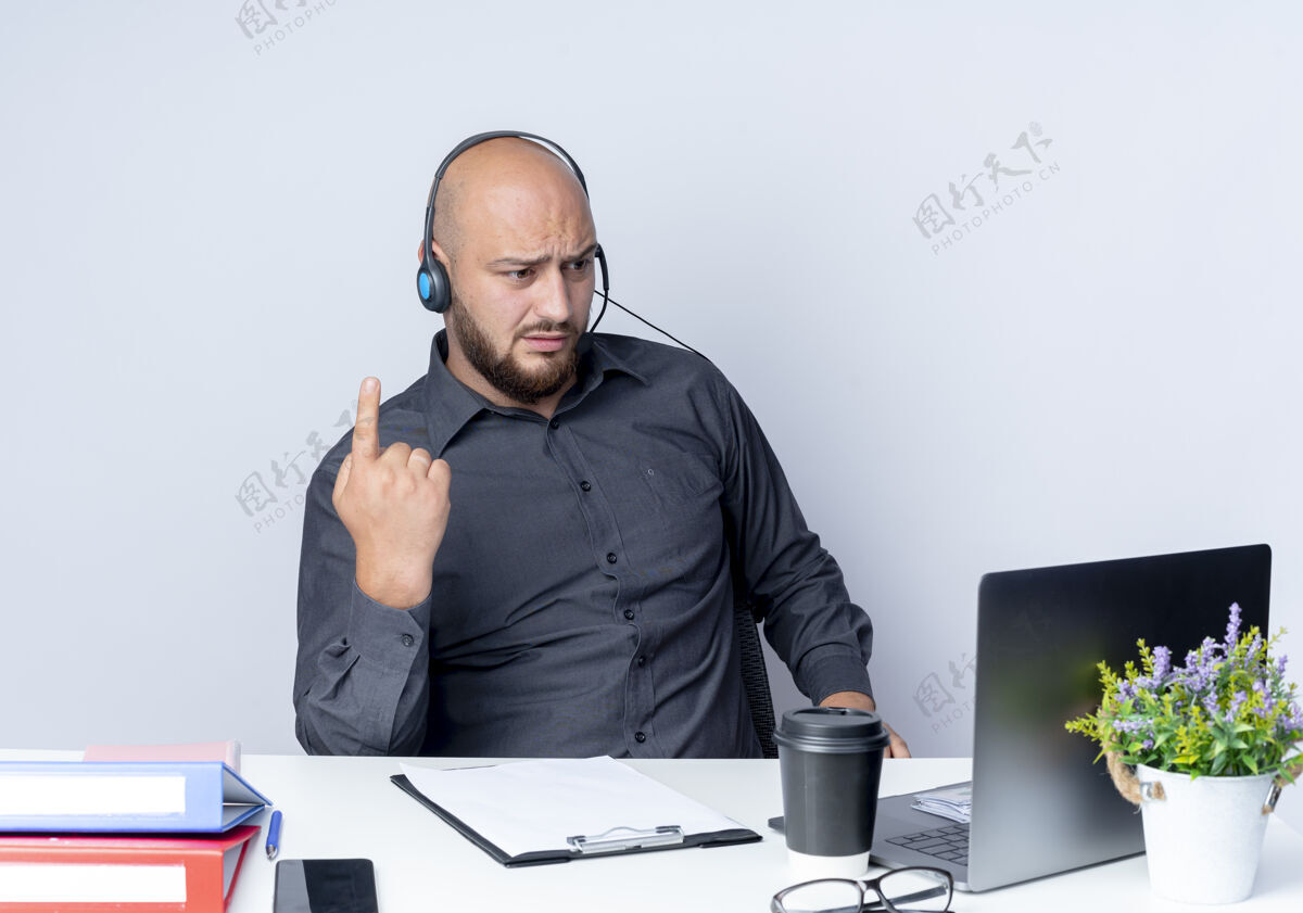 笔记本电脑戴着耳机的秃头呼叫中心年轻人坐在办公桌旁 举着手指 看着隔离在白色背景上的笔记本电脑电话不愉快提高