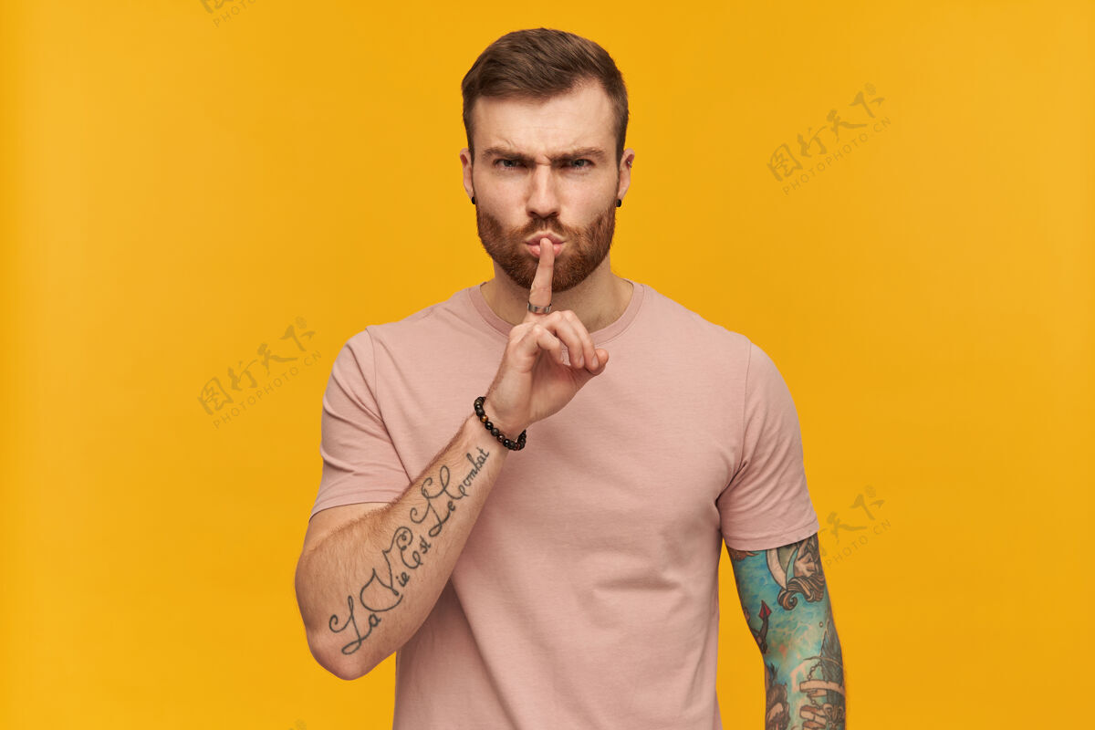 手势愤怒恼怒的年轻人穿着粉色T恤 留着胡子 手上有纹身 看起来很恼火 用手指在黄色的墙上做了个沉默的手势 看着前面镜头谣言墙