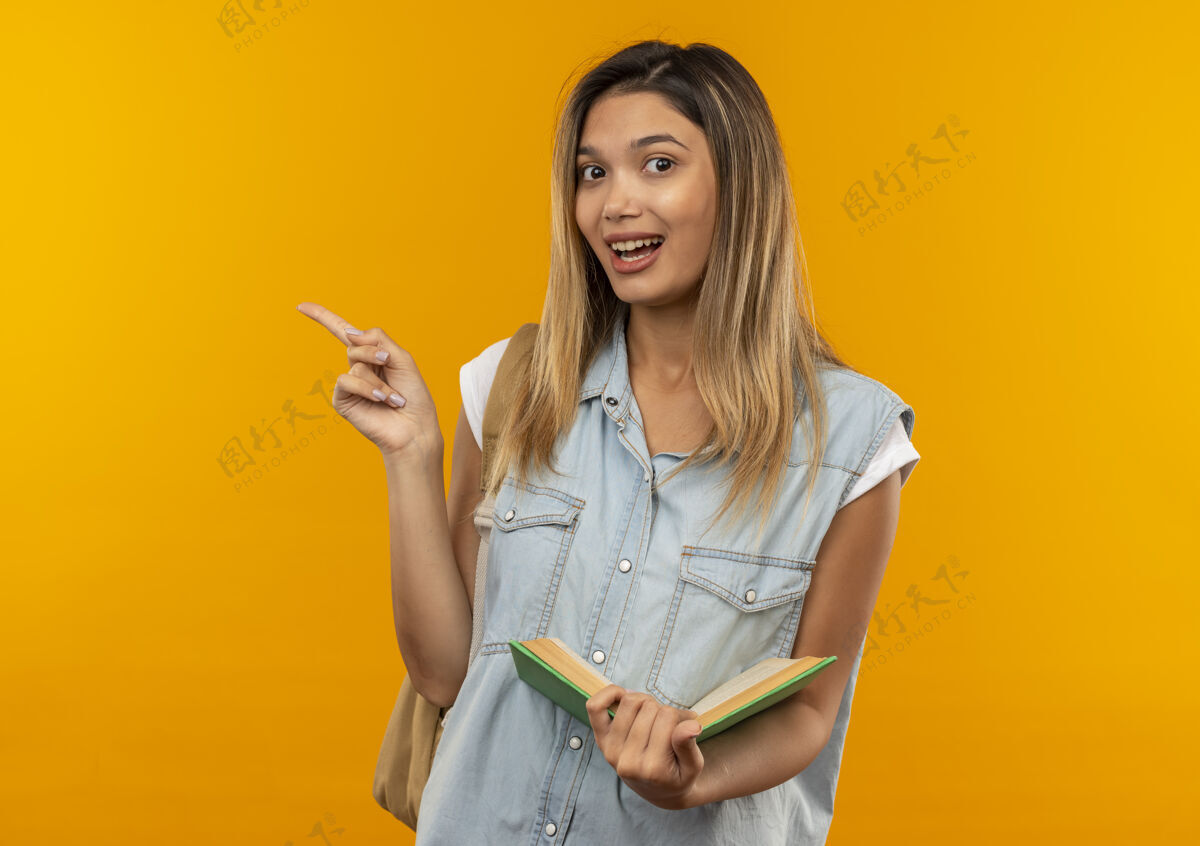 指着快乐的年轻漂亮的女学生 背着书包 拿着打开的书 指着橘子树上孤立的一面袋子快乐橙色