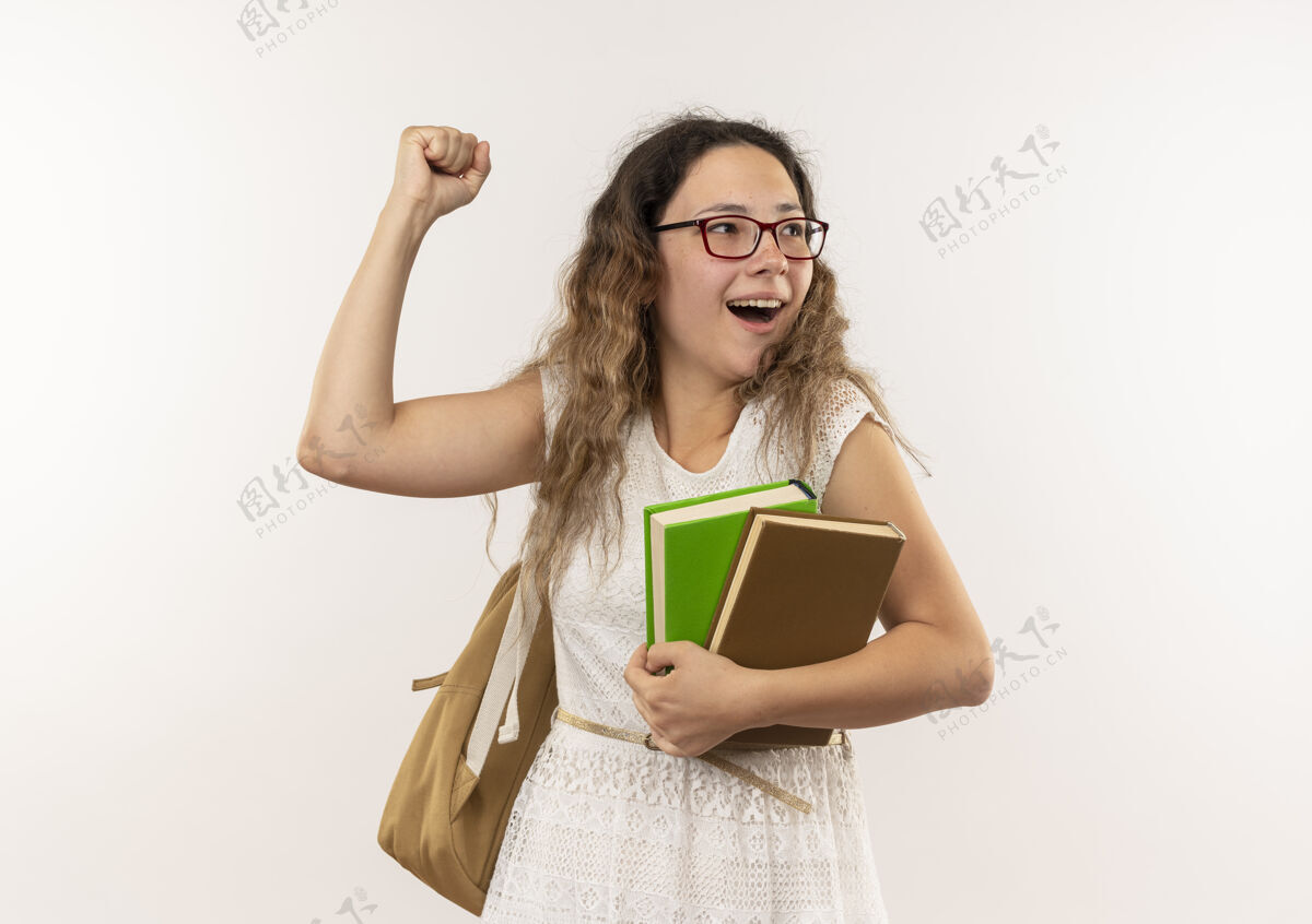 戴上快乐的年轻漂亮的女学生戴着眼镜 背着书包 一边看着书 一边举起拳头孤立在白色的地板上年轻眼镜快乐