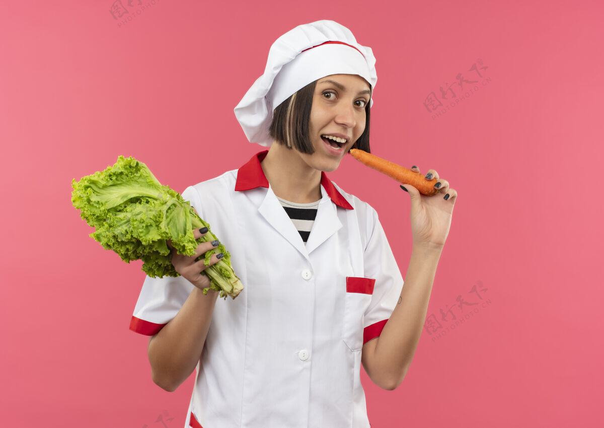 生菜快乐的年轻女厨师身着厨师制服 手里拿着莴苣 试图咬着粉红色的胡萝卜胡萝卜制服年轻