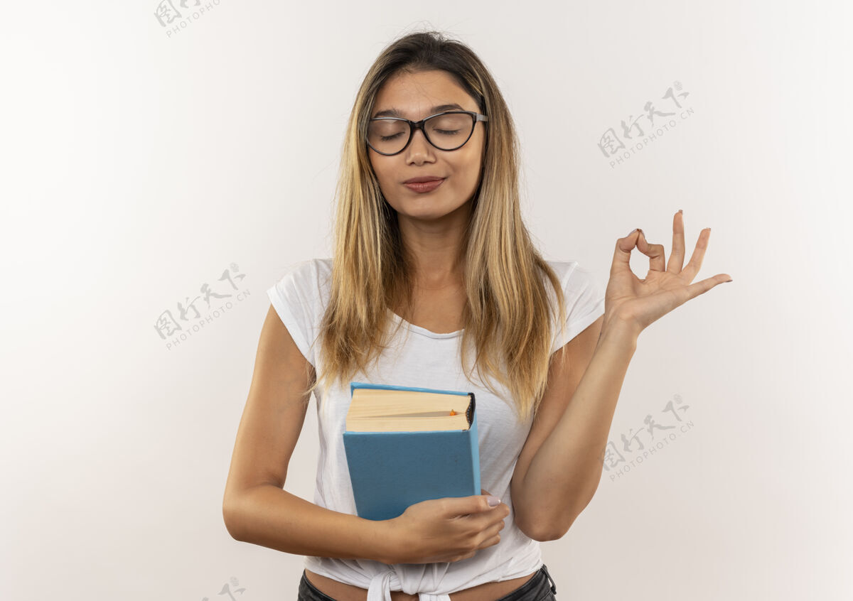 好安静的年轻漂亮的学生女孩戴着眼镜 背着包拿着书 闭着眼睛做着“ok”标志 隔离在白色背景上 留有复印空间戴着年轻背
