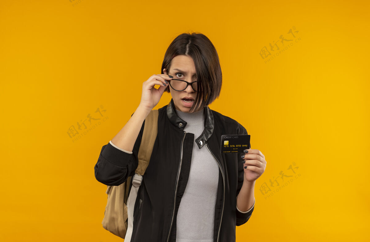 佩戴印象深刻的年轻女学生戴着眼镜背着包拿着信用卡把手放在眼镜上孤立的橙色信用包眼镜