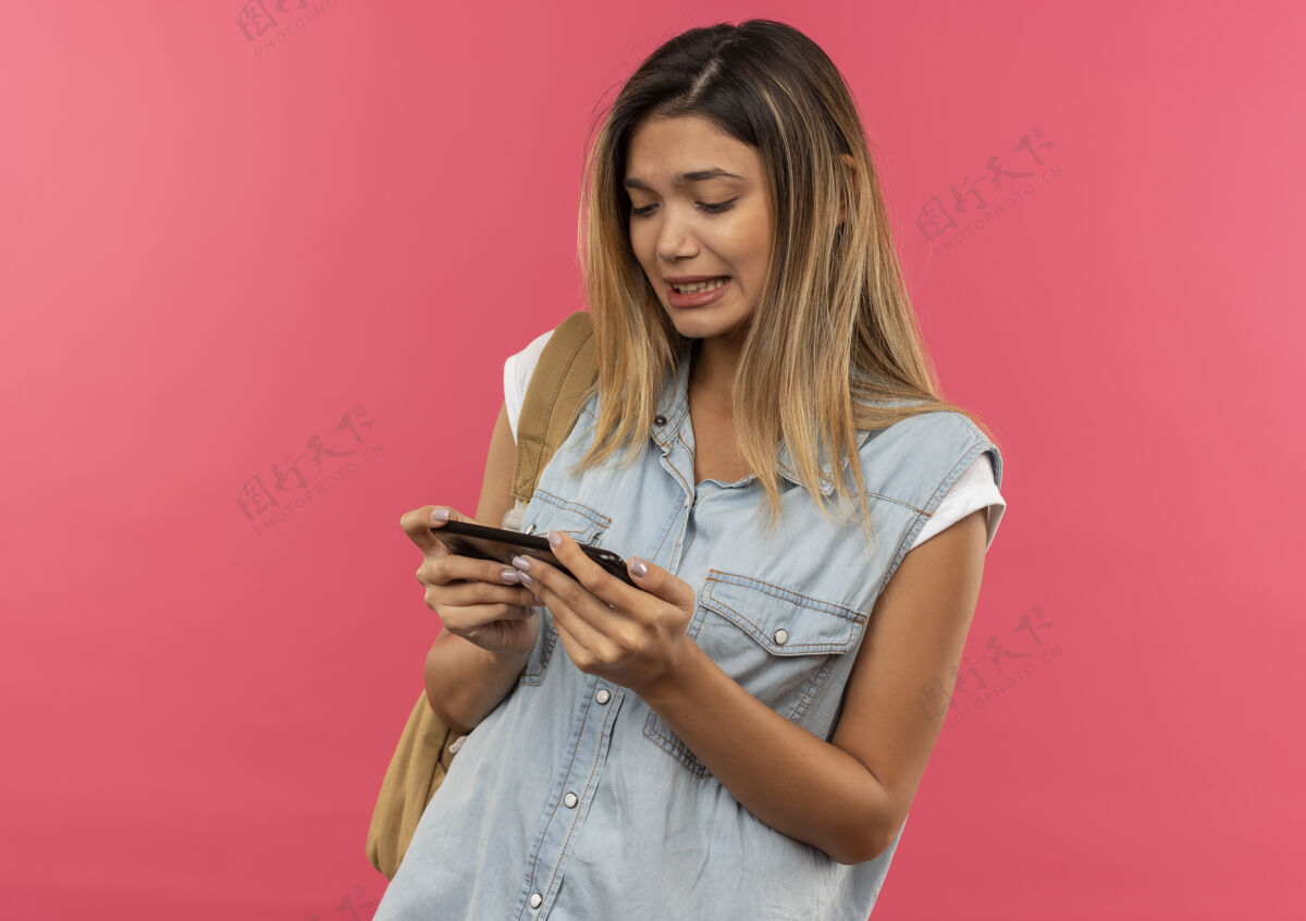 学生未出租的年轻漂亮的学生女孩戴着背包拿着 看着手机孤立的粉红色背景与复制空间女孩包背