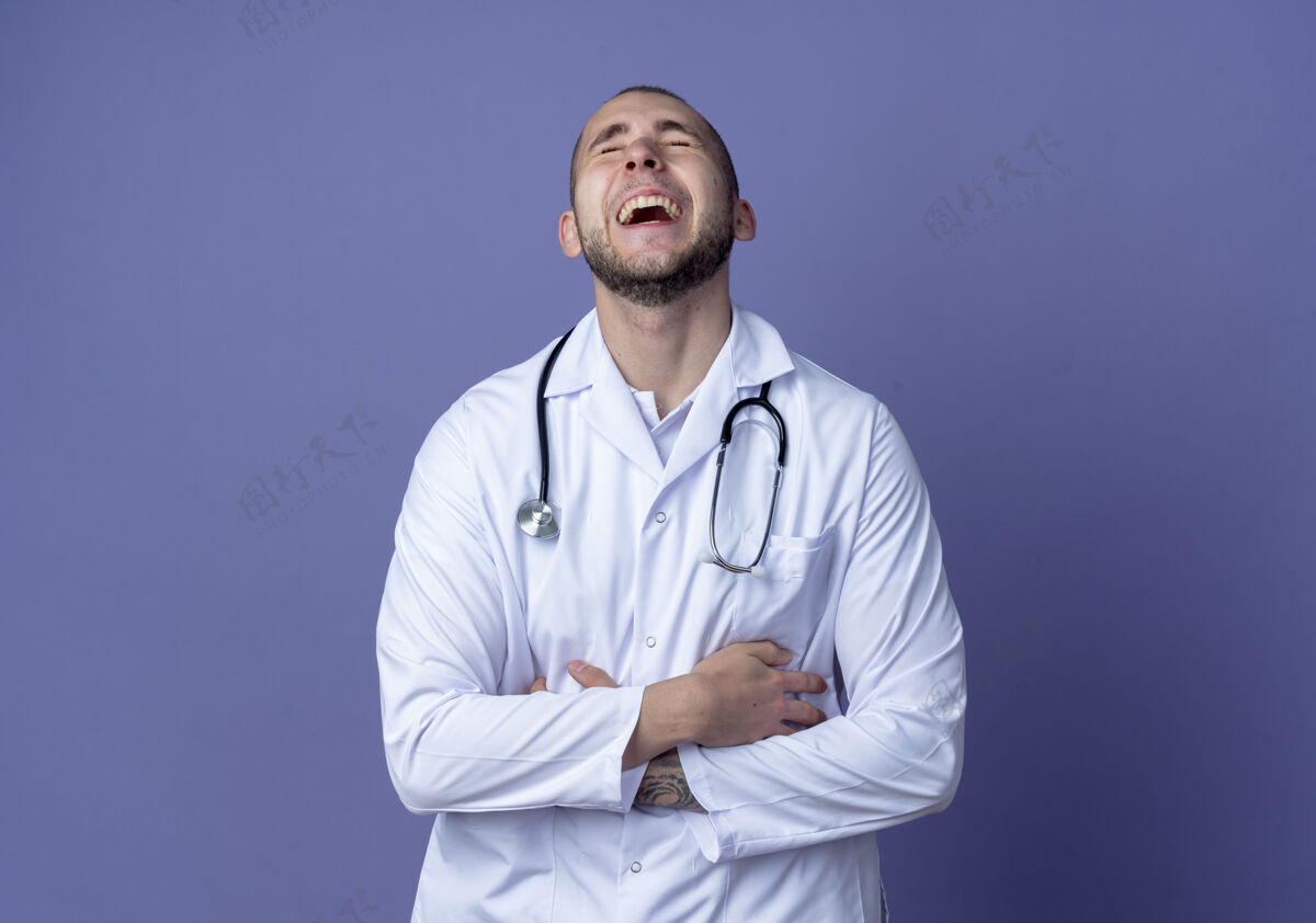 医生笑着的年轻男医生穿着医用长袍和听诊器双手交叉放在肚子上 闭着眼睛隔离在紫色背景上 留有复印空间男性长袍笑