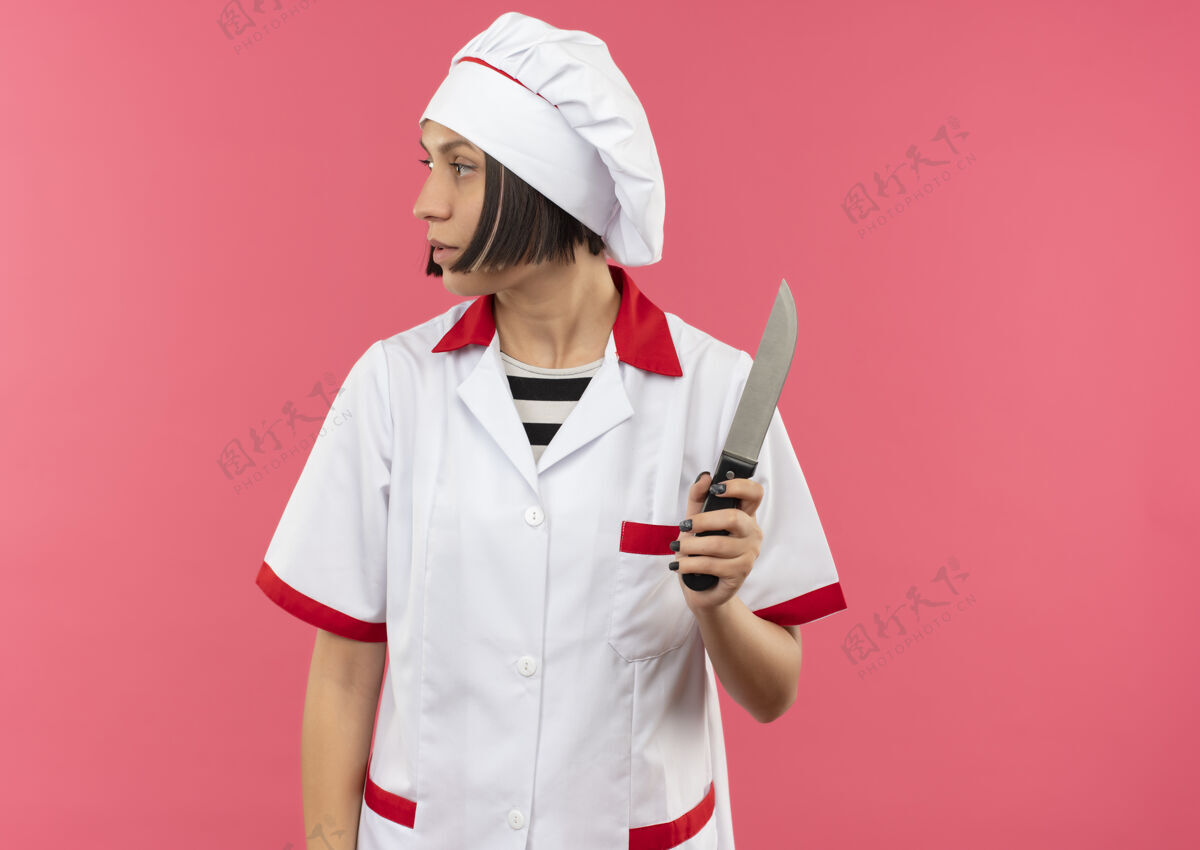 厨师年轻的女厨师身着厨师制服 手持刀 看着旁边的粉红色背景上孤立着的复制空间烹饪壁板制服