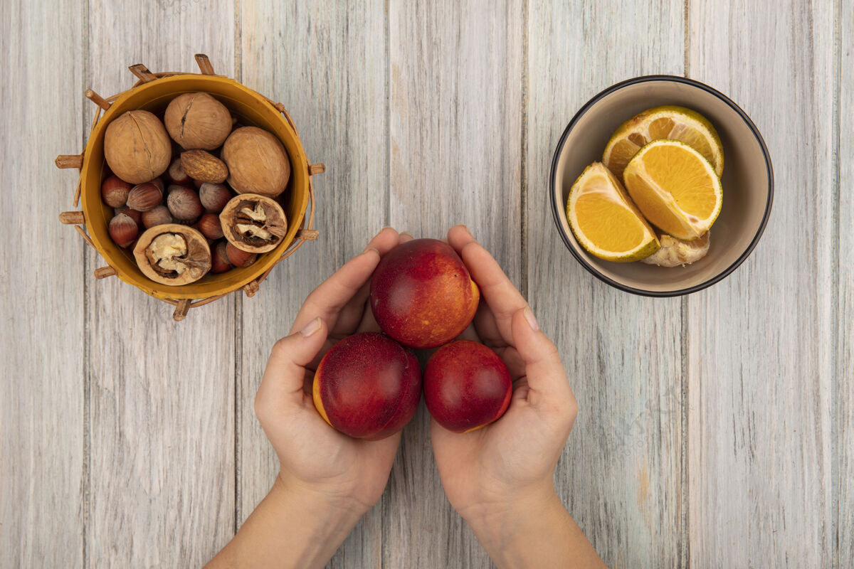 健康顶视图中的女性手拿着新鲜的红色桃子和坚果放在桶上 橘子放在一个灰色木制背景的碗上灰色持有桃子