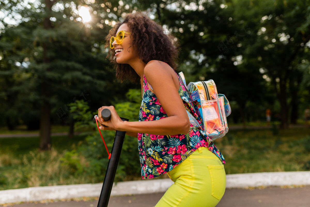 积极年轻时尚的黑人女子在公园里玩得很开心骑着电动脚踏车在夏天的时尚风格 五颜六色的时髦服装 穿着背包和黄色太阳镜埃斯库特车辆环保