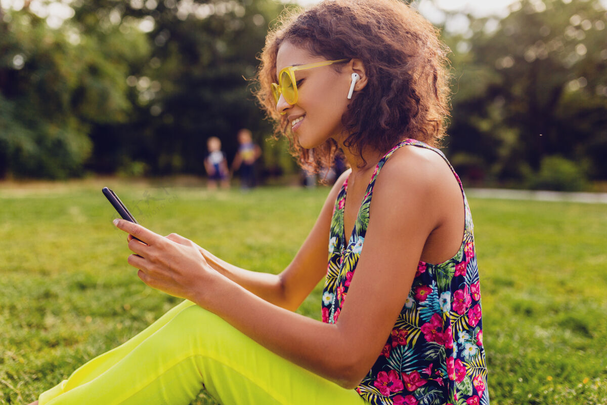 音乐年轻时尚微笑的黑人女子用智能手机听音乐无线耳机在公园里玩得开心 夏日时尚缤纷风格黄色人女士