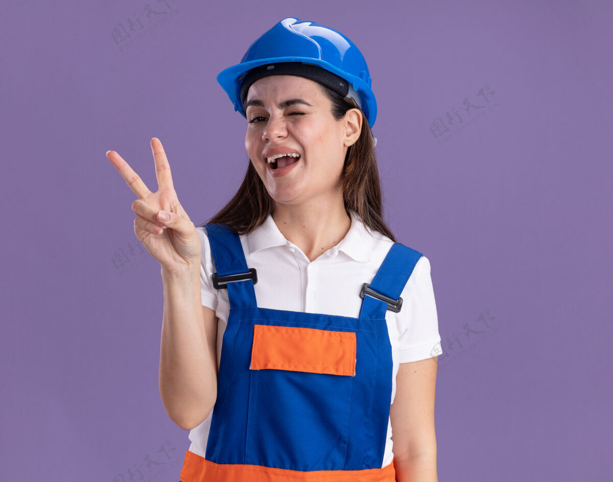 手势在紫色的墙上 一个身穿制服的年轻的建筑工人高兴地眨着眼睛 表现出一种与世隔绝的和平姿态制服眨眼欢乐