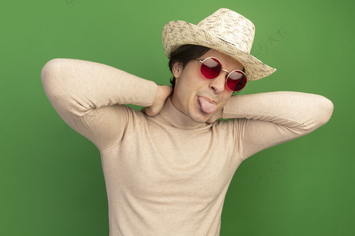 穿快乐的年轻帅哥戴着帽子戴着眼镜露出舌头双手放在脖子上隔离在绿色的墙上年轻人眼镜表演