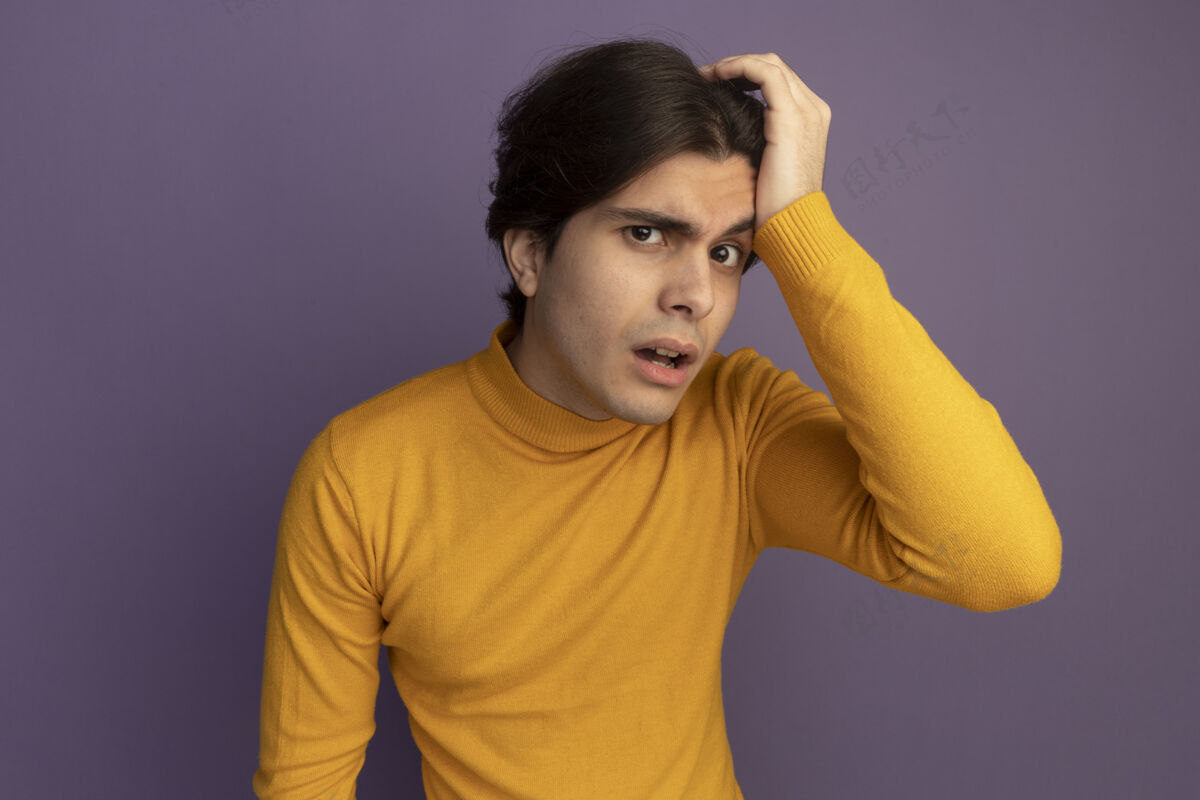 可疑可疑的年轻帅哥穿着黄色高领毛衣 手放在紫色的墙上孤立的头上毛衣头年轻