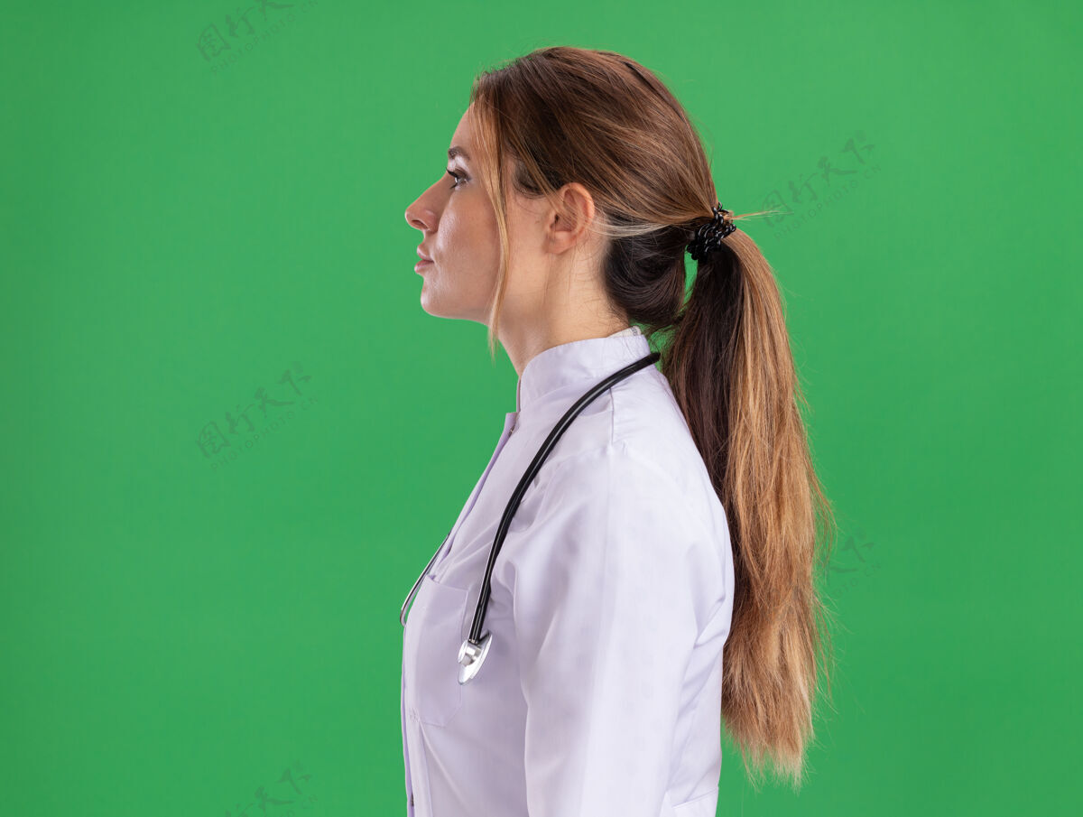 个人资料站在侧视图年轻女医生穿着医用长袍听诊器隔离在绿色墙上长袍听诊器医生