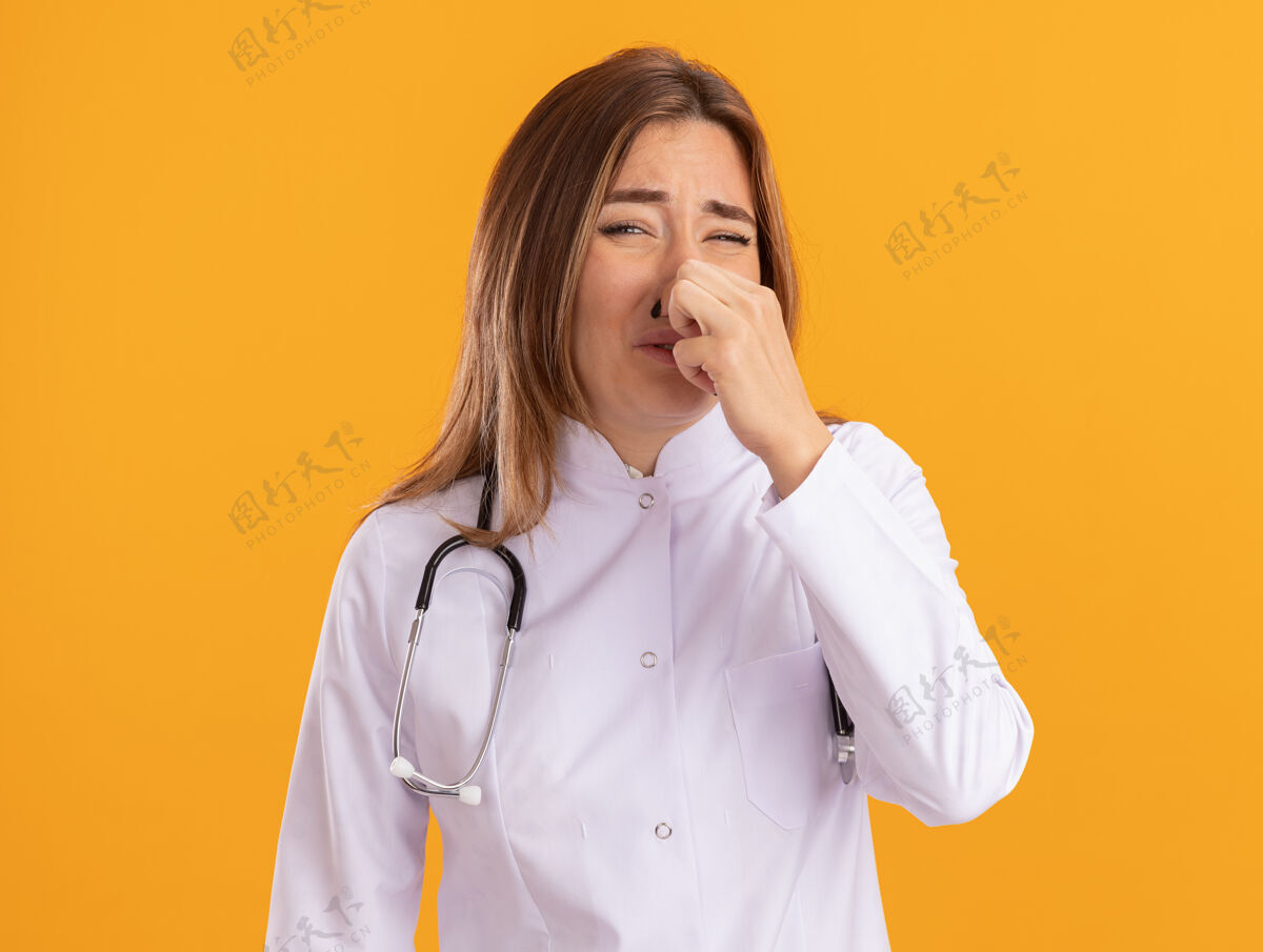 医生一个穿着医用长袍 戴着听诊器 神经质的年轻女医生抓住了隔离在黄色墙上的鼻子年轻医疗吱吱