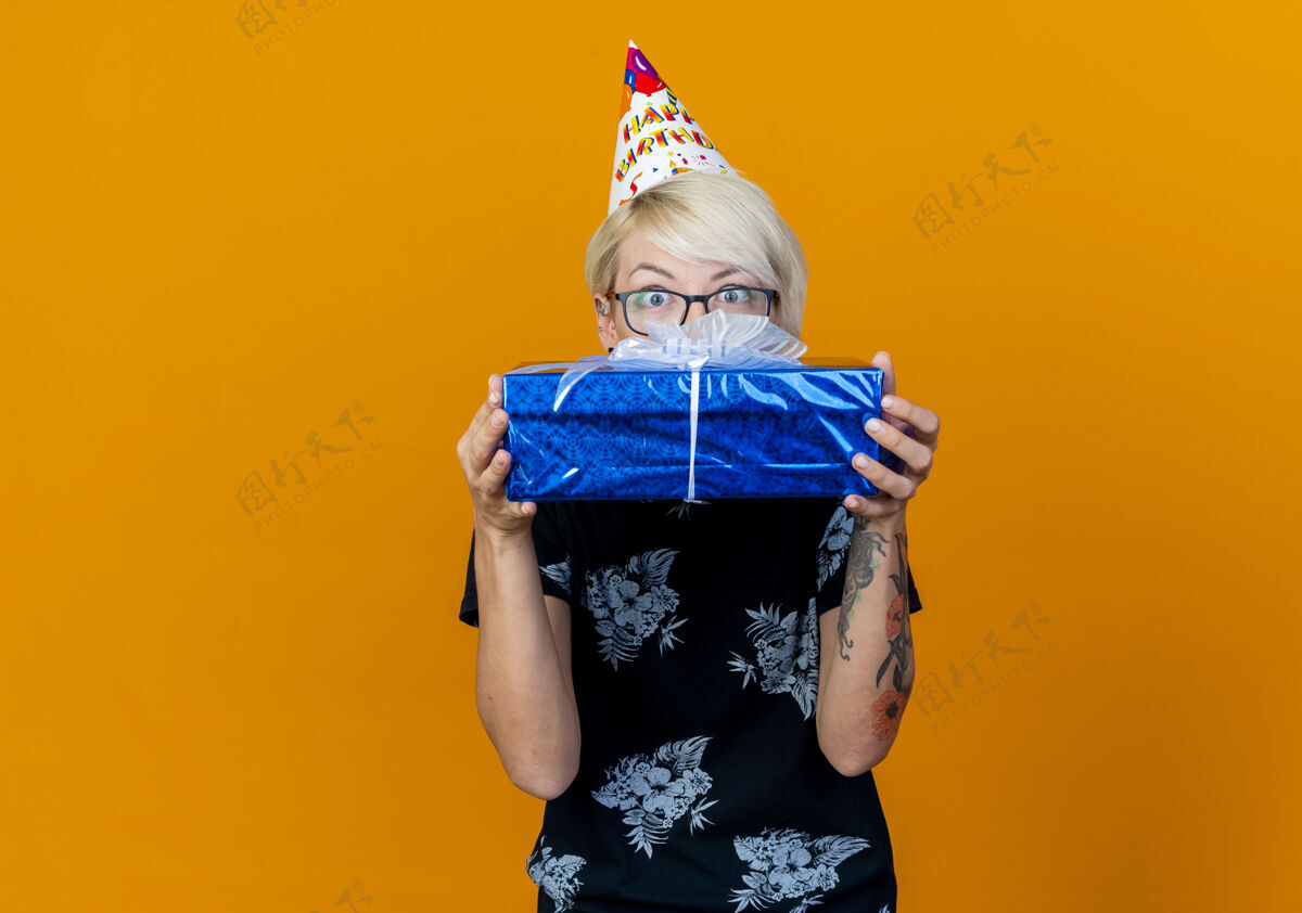 年轻令人印象深刻的年轻金发派对女孩戴着眼镜 戴着生日帽 拿着礼品盒 从后面看着相机 在橙色背景上与复制空间隔离聚会礼物橙色