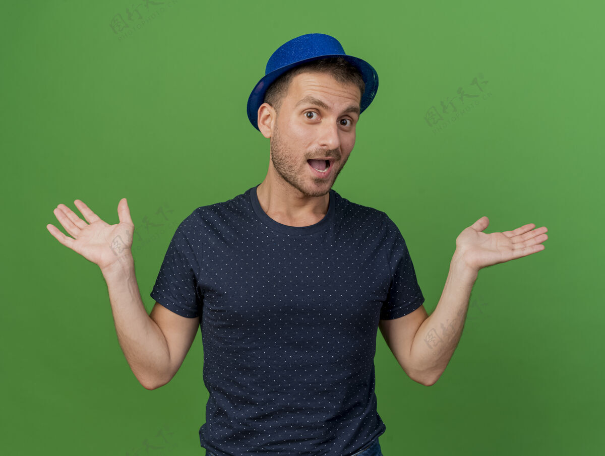 举行快乐英俊的高加索男子戴着蓝色的派对帽 双手张开 在绿色背景上与世隔绝 留有复制空间绿色蓝色人