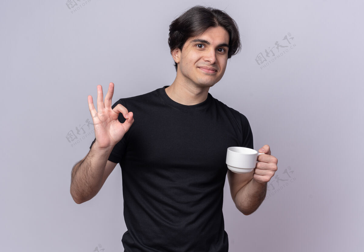 拿着高兴的年轻帅哥穿着黑色t恤 拿着一杯咖啡 在白墙上显示出良好的姿态杯子T恤表演