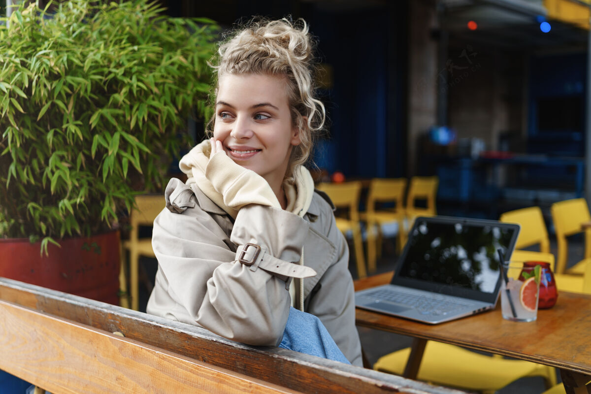 笔记本电脑一个美丽的微笑女孩坐在户外咖啡馆的后视图 看着路人 看着街道 正在休息咖啡馆工作咖啡