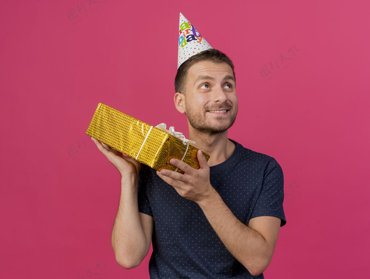 男士戴着生日帽的白种帅哥拿着礼盒 看着粉色背景上孤立的一面 还有复制空间盒子帽子复印件