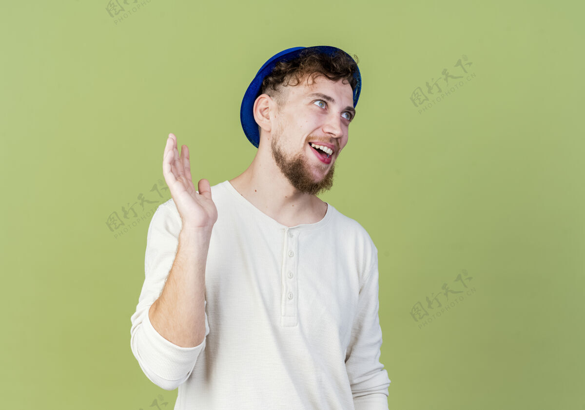 挥手快乐的年轻英俊的斯拉夫党家伙戴着党的帽子看着直挥手在橄榄绿背景与复制空间孤立的人空间斯拉夫人直