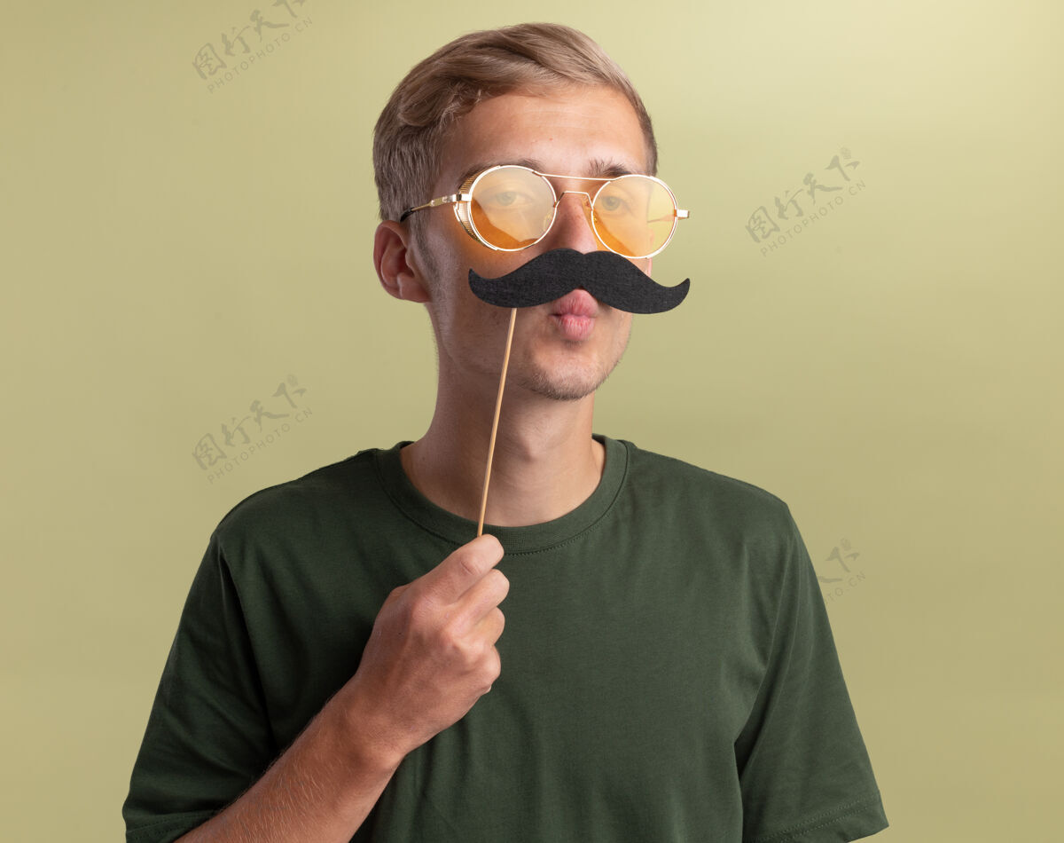假快乐的年轻帅哥穿着绿色衬衫 戴着眼镜 在橄榄绿的墙上贴着假胡子胡子欢乐家伙