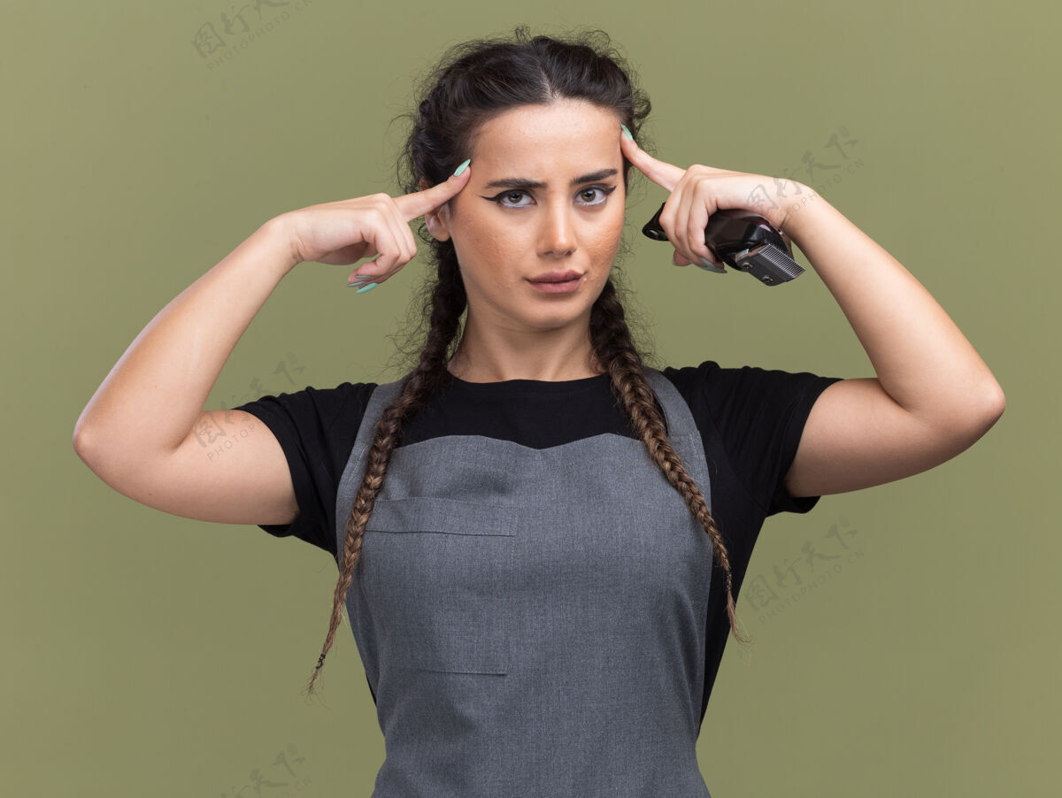 印象给人印象深刻的年轻女理发师穿着制服拿着理发器把手指放在太阳穴上隔离在橄榄绿的墙上橄榄手指剪子