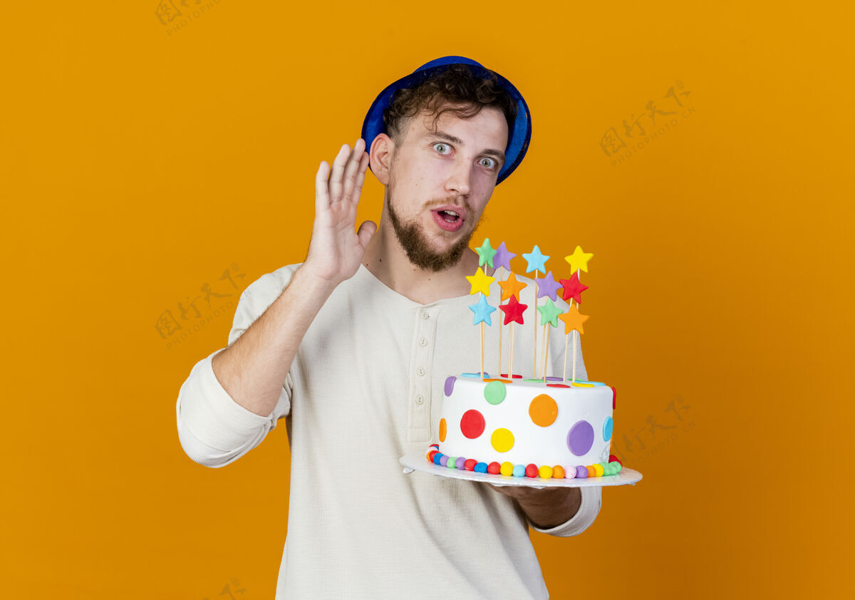 拿着好奇的年轻帅哥斯拉夫人 戴着派对帽 拿着生日蛋糕 拿着星星 看着相机 听不到你的手势 被隔离在橙色背景上 留着复制空间太空男人帽子
