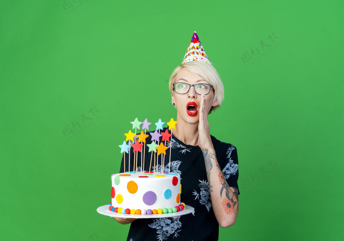 生日令人印象深刻的年轻金发派对女孩戴着眼镜 戴着生日帽 拿着生日蛋糕 星星们一边看着一边窃窃私语 在绿色背景上与复制空间隔离帽子耳语空间