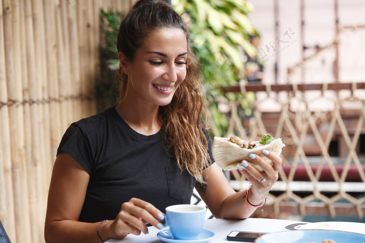 人皮肤黝黑的健康女人 穿着t恤坐在咖啡厅露台上 吃着早餐 喝着咖啡咖啡热带休闲