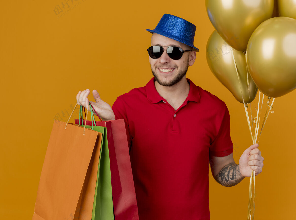 橘子快乐的年轻帅哥斯拉夫党家伙戴着墨镜和党帽子举行气球和纸袋孤立的橙色背景年轻人抱着气球