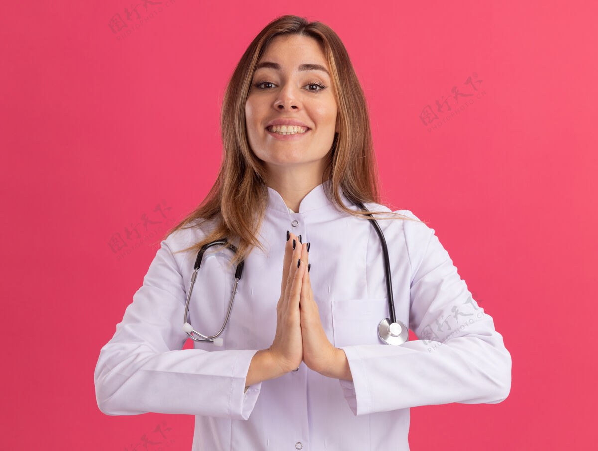 年轻高兴的年轻女医生穿着医用长袍 听诊器显示祈祷手势隔离在粉红色的墙上祈祷女请