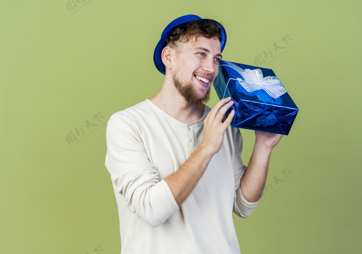 欢乐快乐的年轻帅哥斯拉夫党家伙戴着党的帽子看起来直举行礼盒孤立的橄榄绿背景与复制空间直空间盒子