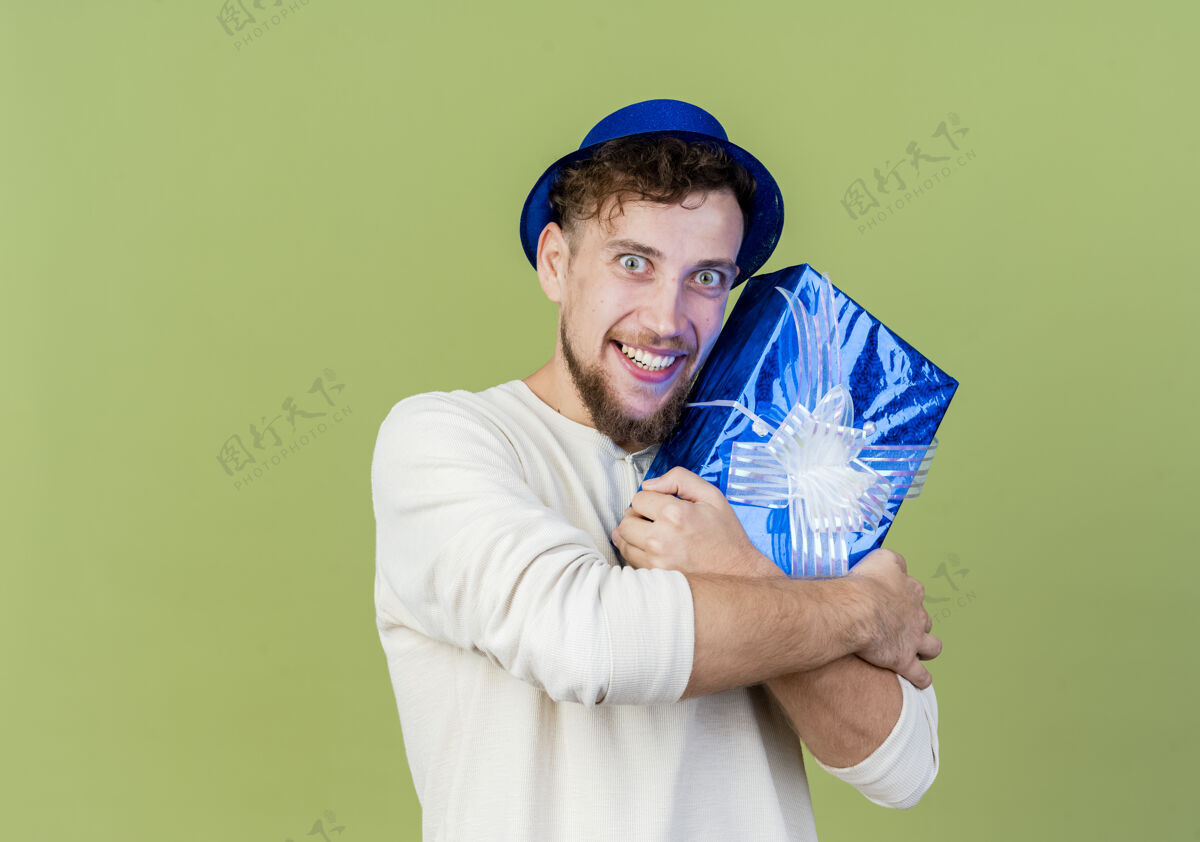 空间微笑的年轻帅哥斯拉夫党家伙戴着党帽子拿着礼品盒看着相机孤立的橄榄绿背景与复制空间小伙子微笑盒子