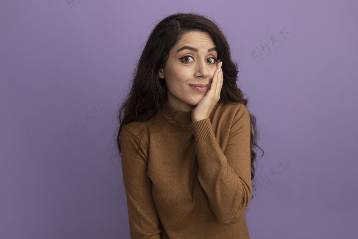 不舒服穿着棕色高领毛衣 手放在脸颊上的年轻漂亮女孩被隔离在紫色的墙上高领毛衣穿着年轻