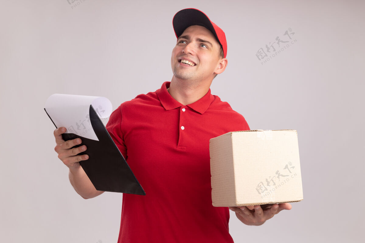 男人微笑着抬头看年轻的送货员 穿着制服 帽子夹着盒子 剪贴板被隔离在白色的墙上帽子年轻人戴着
