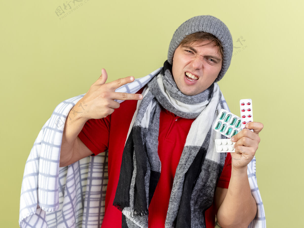 包装自信的年轻英俊的金发病夫戴着冬天的帽子 围巾裹着格子布 手里拿着几包药丸 看着橄榄绿背景上孤立的摄像机围巾年轻帽子