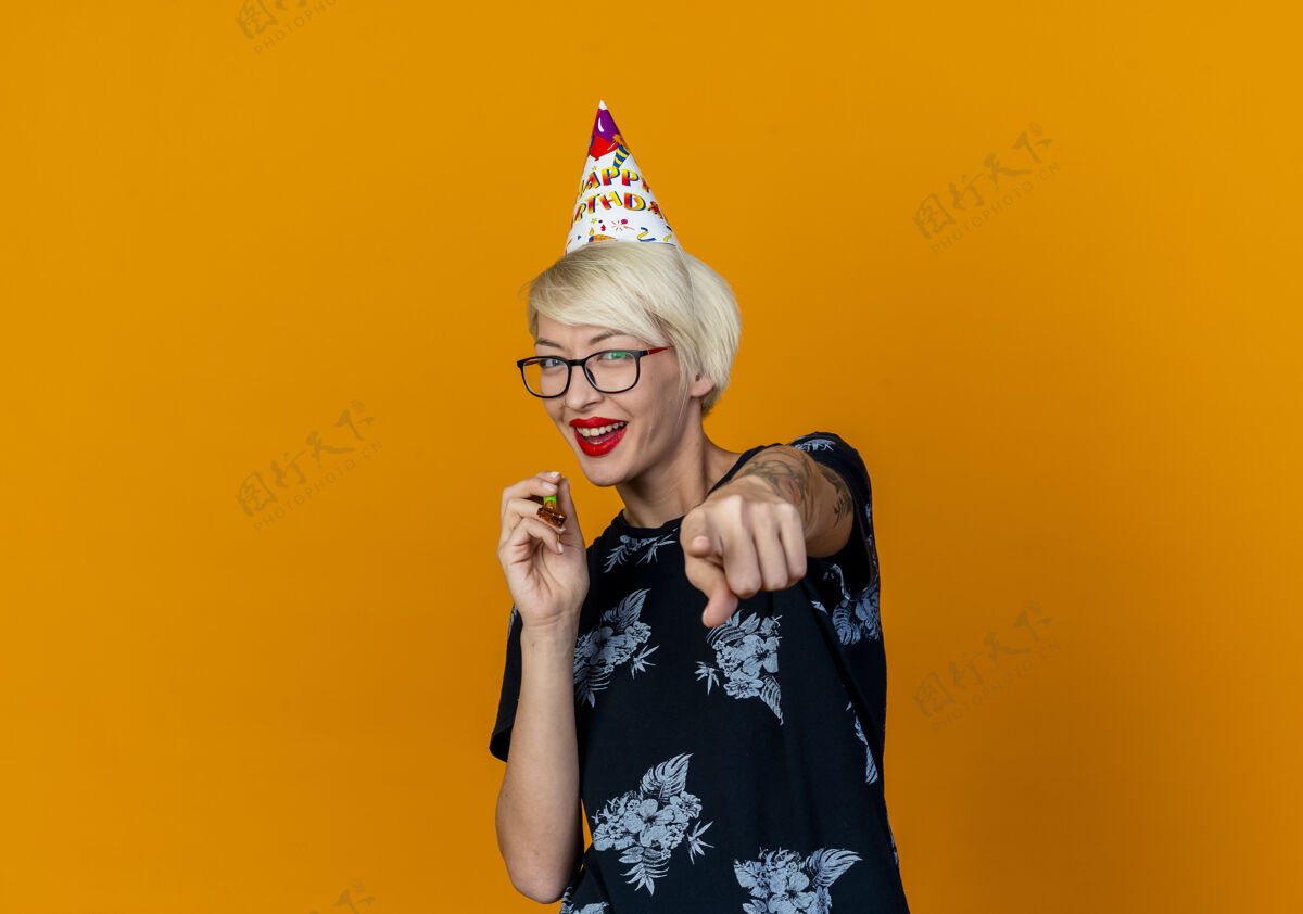 空间快乐的年轻金发派对女孩戴着眼镜 戴着生日帽 手持派对吹风机 看着并指着隔离在橙色背景和复印空间的相机女孩帽子金发