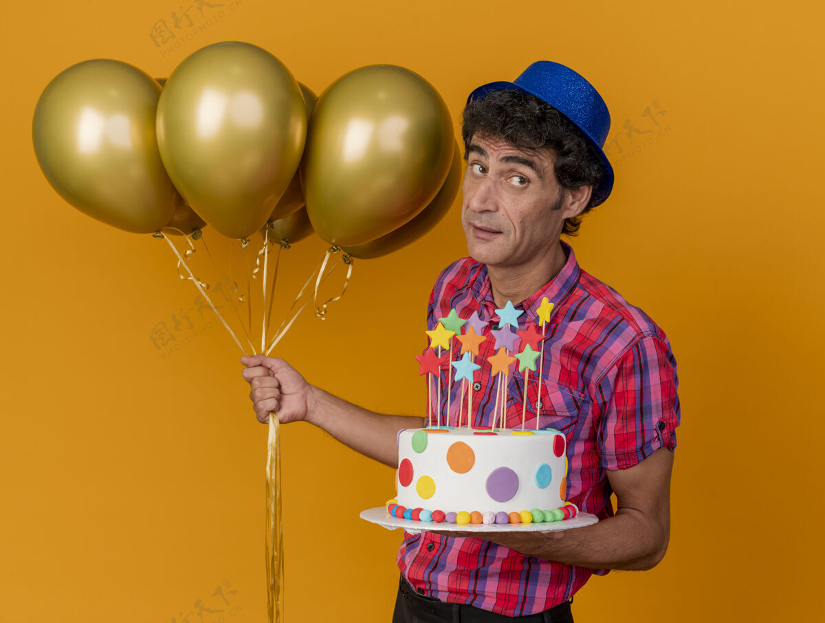 视图印象深刻的中年白种人党的人戴着党的帽子站在侧视图举行气球和生日蛋糕看着橙色背景上孤立的一面个人资料帽子男人