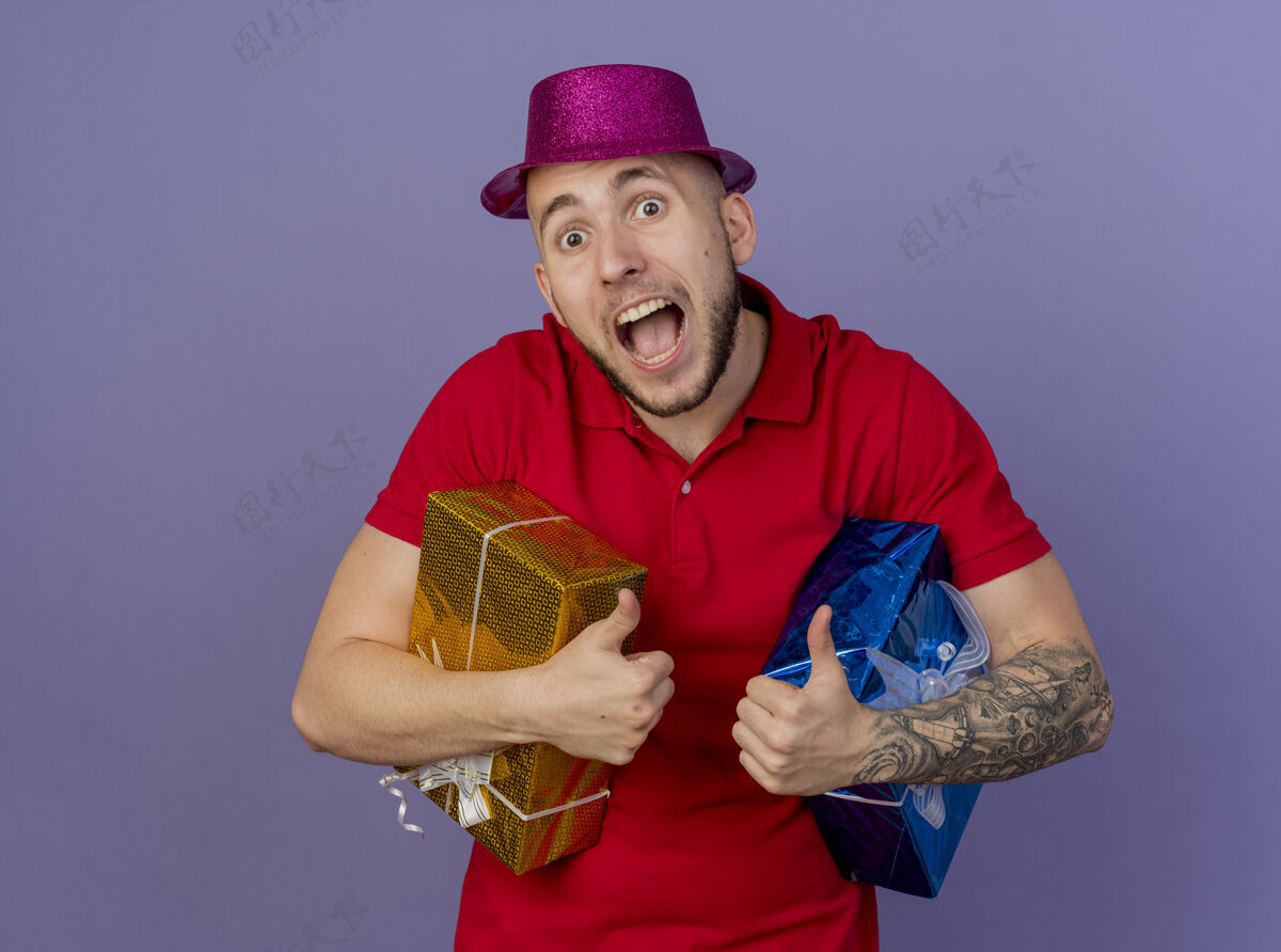 派对令人印象深刻的年轻英俊的斯拉夫党的家伙戴着党的帽子 手持礼品包 看着在紫色背景与复制空间隔离相机帽子斯拉夫人紫色