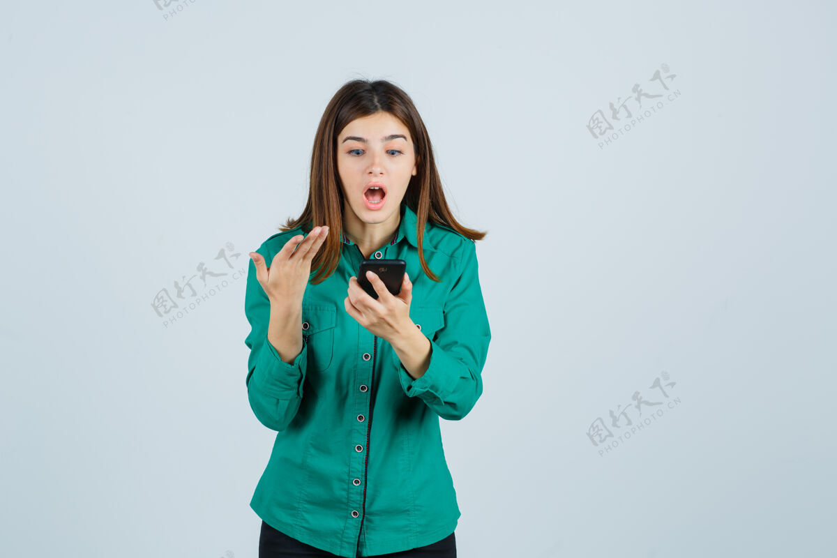 年轻年轻女孩看着手机 穿着绿色上衣 黑色裤子 惊讶地把手伸向手机 看上去很震惊 正对着手机成人面部女性