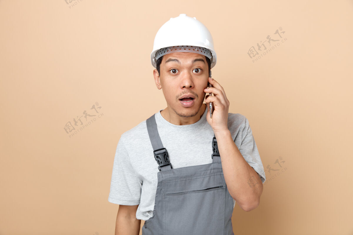 奶油惊讶的年轻建筑工人戴着安全帽和制服在打电话穿惊喜电话