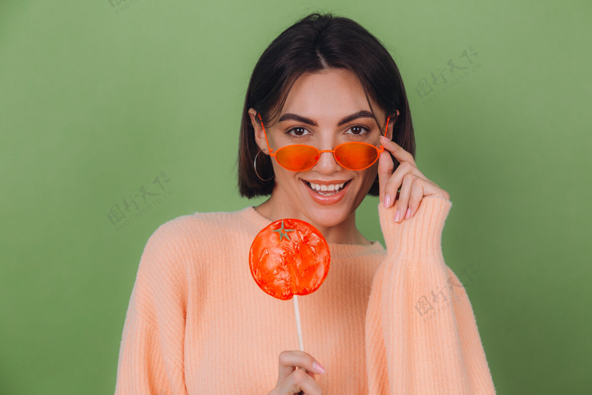 魅力年轻时尚的女士穿着休闲的桃色毛衣 戴着橙色的眼镜 隔离在绿色橄榄色的墙上 带着橙色棒棒糖的正面微笑复制空间棒棒糖太阳镜青年