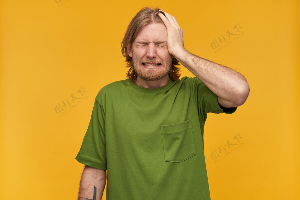 休闲紧张的男人 后悔的家伙金发 胡子和胡子穿着绿色t恤有纹身把手掌放在他的头上感到疼痛站在黄色的墙隔离手掌胡须室内