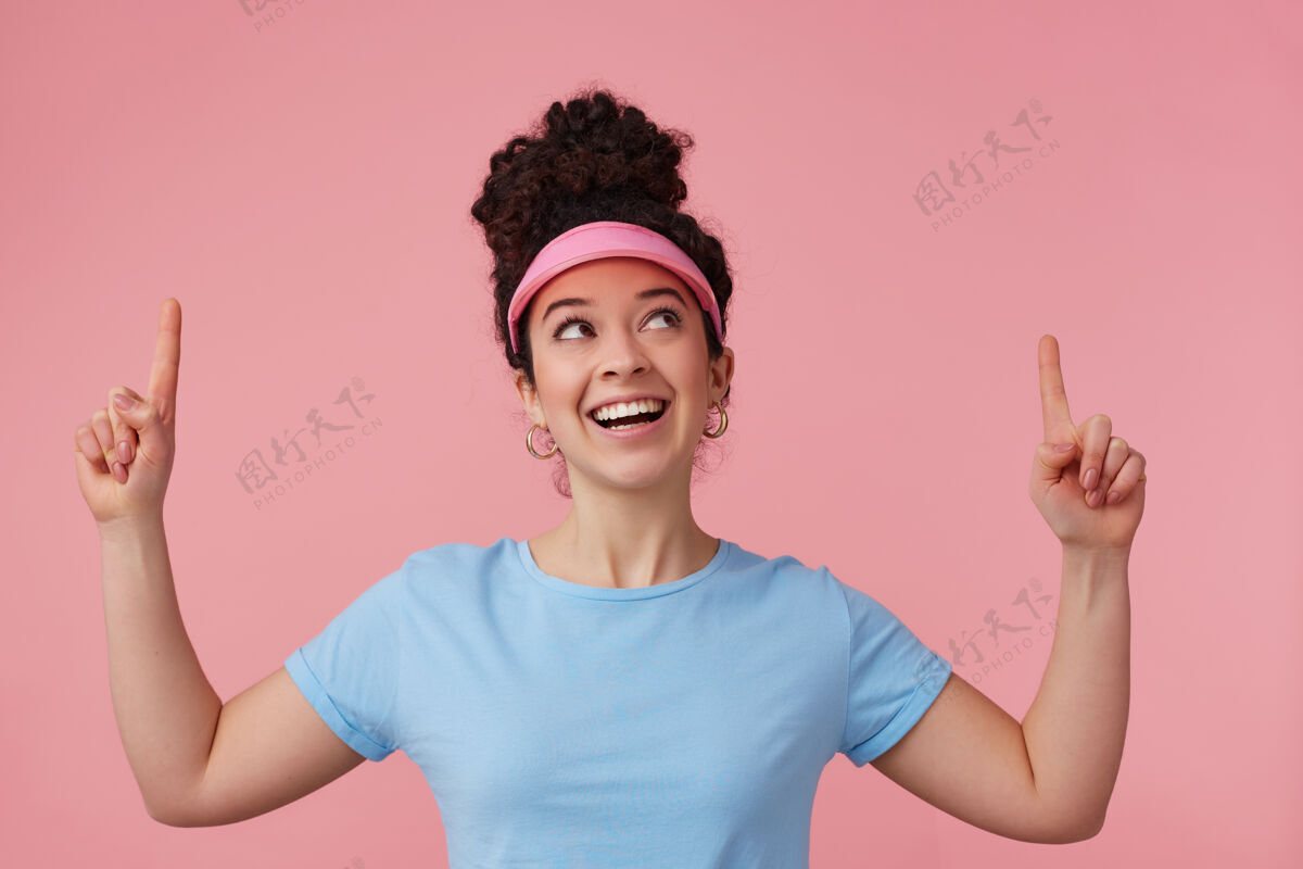 游戏俏皮的女人 漂亮的女孩 深色卷发 戴着粉红色的帽舌 耳环和蓝色的t恤已经化妆了服装拍摄T恤