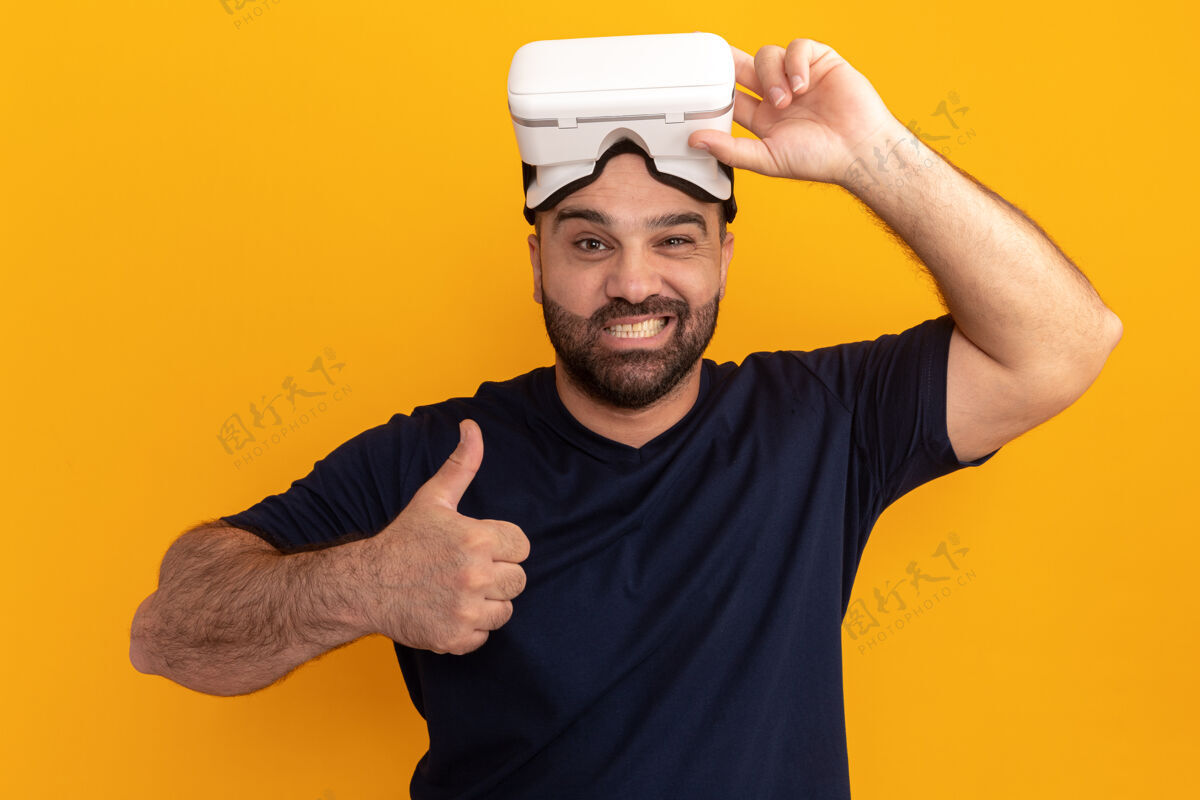 男人留着胡子的男人穿着海军t恤 戴着虚拟现实的眼镜 快乐而积极的微笑 站在橙色的墙上竖起大拇指虚拟的大拇指胡须